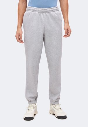 Lichtgrijze joggingbroek met elastische tailleband en boorden gedragen met witte sneakers bij een persoon die staat tegen een effen achtergrond.