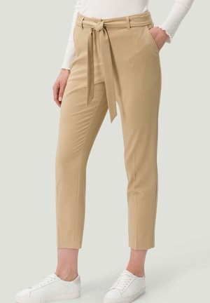 Femme portant un pantalon beige fuselé avec une ceinture nouée à l'avant, un haut blanc à manches longues et des baskets blanches, debout les mains dans les poches.