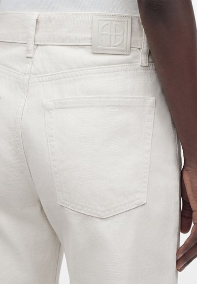 Gros plan d'une personne portant un jean denim blanc cassé montrant une poche arrière et un patch logo embossé sur la ceinture.