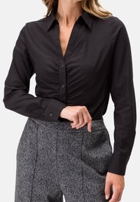 Blouse noire à manches longues avec un col, présentant un devant froncé et une fermeture à boutons. Portée avec un pantalon à motif pied-de-poule gris.