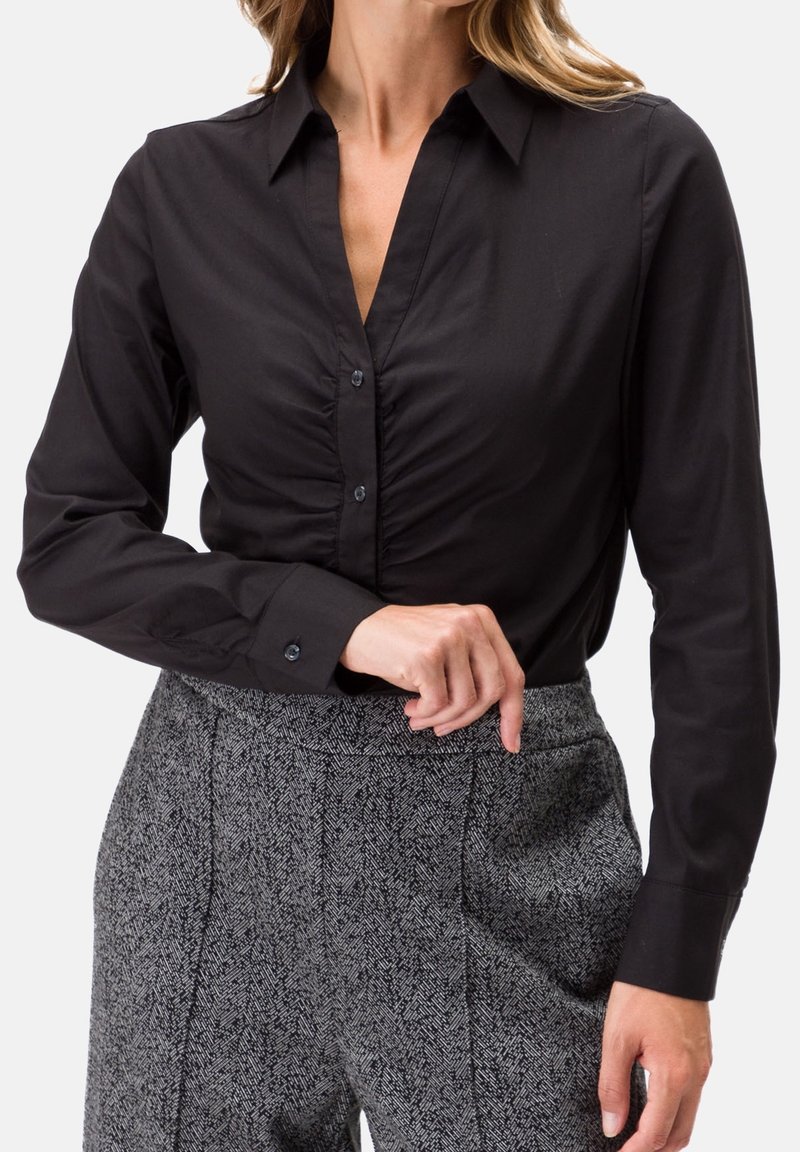 Blouse noire à manches longues avec un col, présentant un devant froncé et une fermeture à boutons. Portée avec un pantalon à motif pied-de-poule gris.