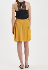 Czarny top na ramiączkach z koronkowym tyłem, połączony z musztardową spódnicą do kolan. Modelka nosi płaskie sandały. Miękka, zwiewna tkanina.