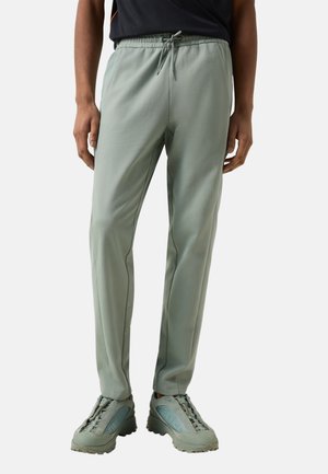 Mann trägt hellgraue schmale Hose mit Kordelzug an der Taille und passende hellgraue Schnür-Trainingsschuhe, stehend.