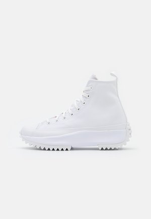 Witte hoge sneaker met dikke zigzagzool, veters aan de voorkant en een lus bij de hiel, tegen een effen lichte achtergrond.