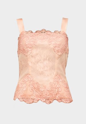 Femme debout portant un caraco en satin rose clair avec des détails en dentelle et un pantalon large en satin assorti sur un fond uni.