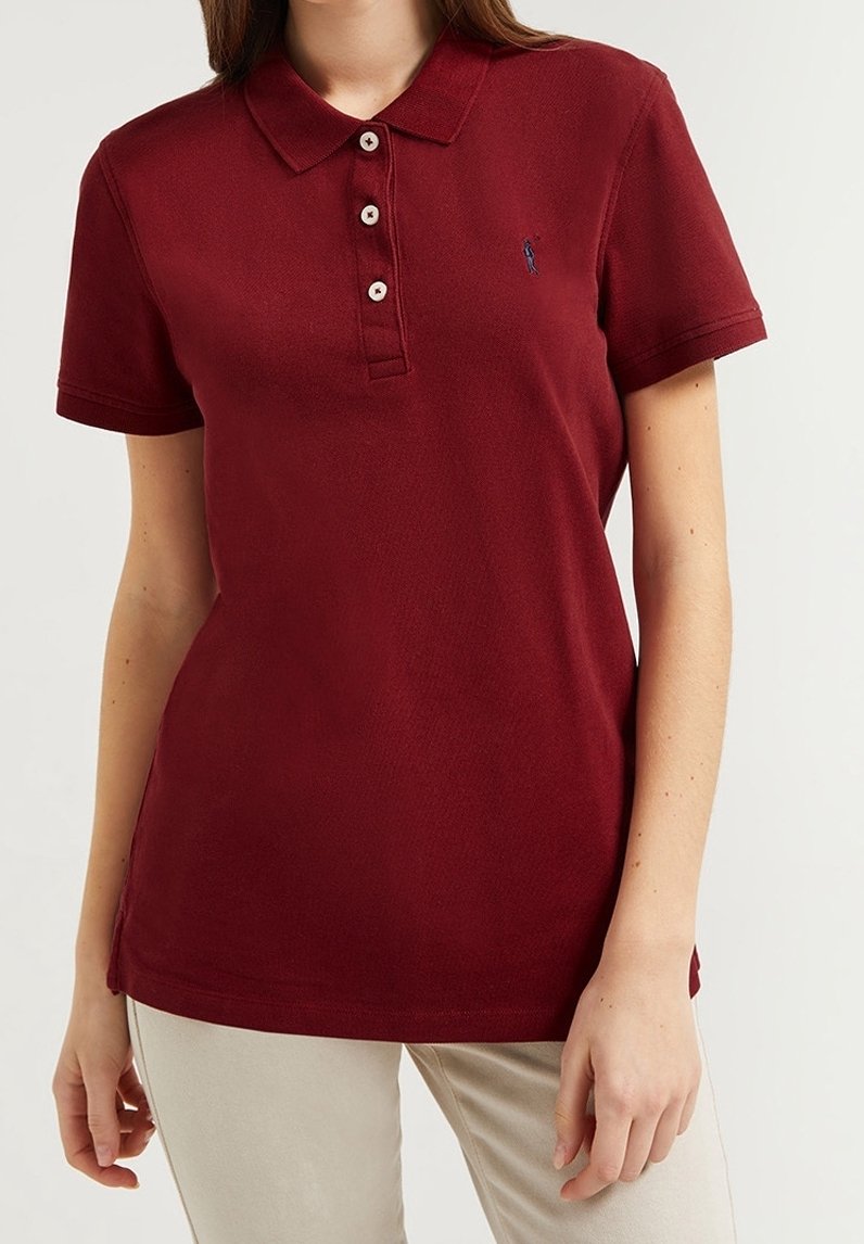 Femme portant un polo bordeaux à manches courtes avec trois boutons et un pantalon beige sur un fond uni.