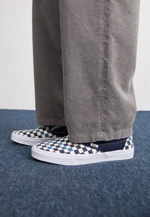 Canvas slip-on sko har et blå og creme ternet mønster med hvid sål; båret med grå kordfløjl bukser.