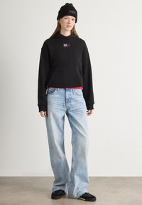Czarna skrócona bluza z kapturem z kieszenią z przodu, czerwona koszulka na warstwy pod spodem, jasnoniebieskie jeansy z szerokimi nogawkami i czarne sneakersy. Gładkie tło.