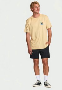 Homem com uma T-shirt bege com gráfico tropical, calções pretos, meias brancas e ténis pretos, de pé com uma mão no bolso, olhando para a direita.