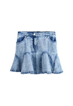 Meisje met lang blond haar draagt een wit AllSaints T-shirt en een blauwe uitlopende spijkerrok, staand tegen een effen grijze achtergrond.