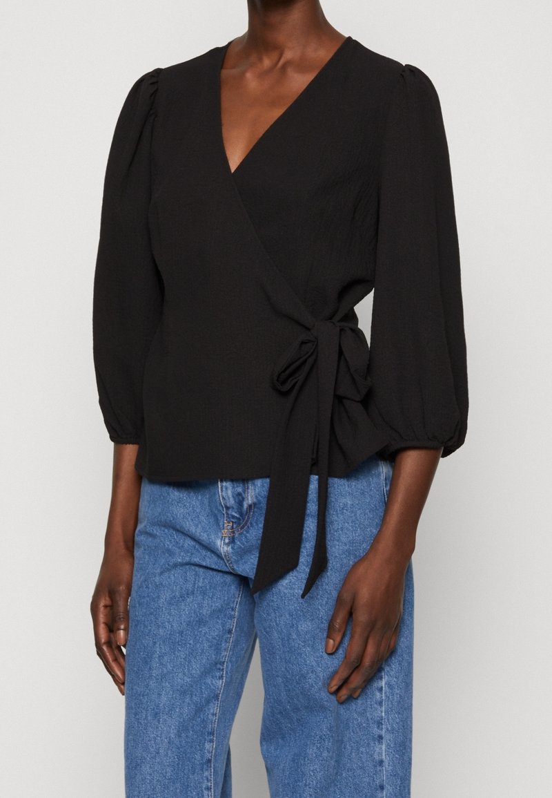 Sort wrap-top med puffede ærmer, en bindedetalje i taljen og et struktureret stof, parret med lyseblå højtaljede jeans.