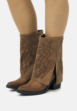 Bottes ankle en suede marron avec un bout pointu, ornées de motifs de vagues brodés et d'une fente sur le côté, reposant sur un talon bloc noir.