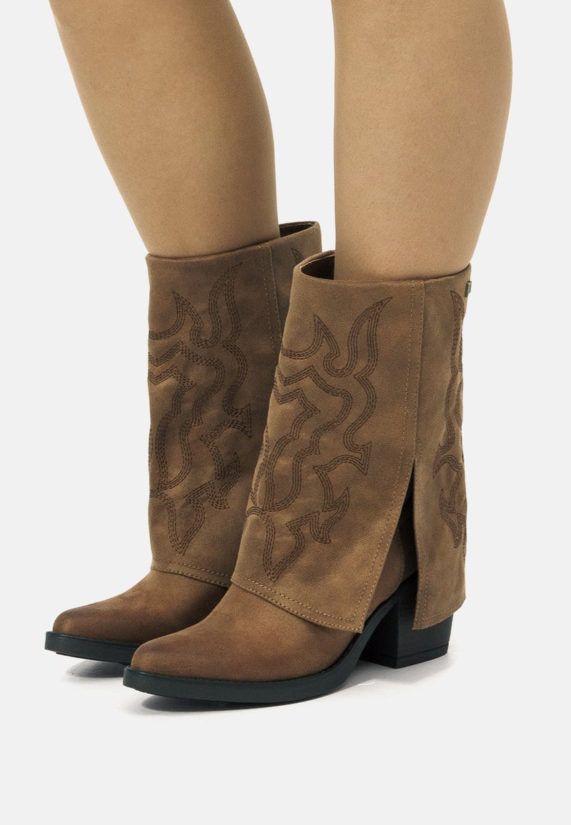 Bottes ankle en suede marron avec un bout pointu, ornées de motifs de vagues brodés et d'une fente sur le côté, reposant sur un talon bloc noir.