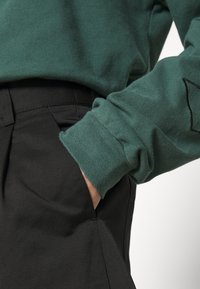 Main d'une personne dans la poche d'un pantalon noir, portant un haut à manches longues vert avec un revers visible et une broderie partielle sur la manche.