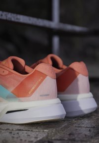 Para różowo-pomarańczowych butów do biegania Adidas Adizero Boston 13 spoczywających na kamiennej nawierzchni z rozmytym ciemnym tłem.