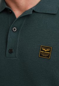 Polo verde texturizado con cuello, dos botones negros y una etiqueta amarilla y negra de PME Legend en el lado izquierdo del pecho.