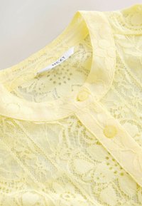 Dzeltena mežģīņu blūze ar apkakli, pogu aizdari un ziedu rakstu. Audums ir viegls un caurspīdīgs, akcentējot smalkas detaļas.