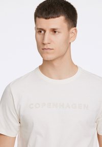 Vaalean kermanvärinen puuvillapaita, jossa on pyöreä kaula-aukko, lyhyet hihat ja pehmeissä väreissä kohokuvioitu teksti "COPENHAGEN LINDBERGH".