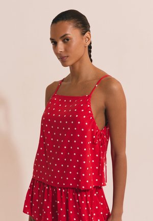 Mujer con el cabello trenzado vistiendo una blusa sin mangas roja con lunares y una falda a juego sobre un fondo liso.