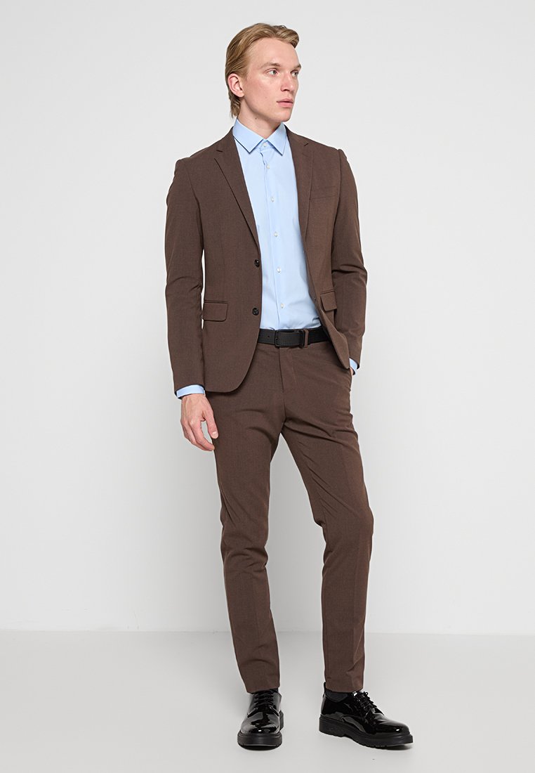 Completo marrone su misura con rever a punta, chiusura con bottone e due tasche. Camicia azzurra chiara sotto. Scarpe nere lucide. Vestibilità slim.