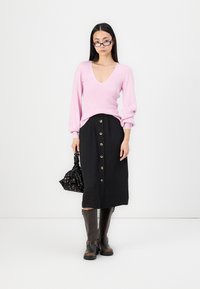 Suéter rosa claro canelado com decote em V e mangas longas. Saia midi preta com botões na frente. Botas castanhas até ao joelho e uma bolsa preta.