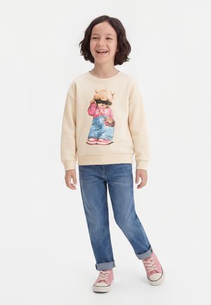 Uśmiechnięte dziecko o średniej długości brązowych włosach nosi beżową bluzę z grafiką misia, niebieskie podwinięte jeansy i różowe trampki.