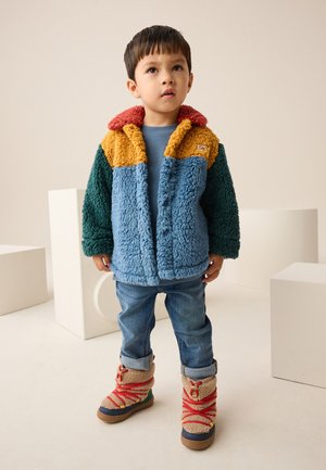 Junger Junge steht drinnen, trägt eine mehrfarbige Fleecejacke, blaue Jeans und beige Stiefel mit roten Schnürsenkeln, blickt leicht nach oben.