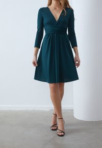 Tealfärgad klänning med djup V-ringning, trekvartsärmar och insamlad midja, som flödar mjukt ner till knä längd. Bärs med svarta remsandaler.