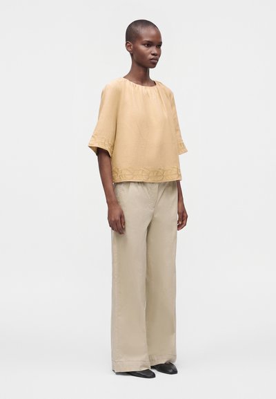 Femme debout portant un haut beige ample avec ourlet brodé et un pantalon crème à jambes larges avec des chaussures noires sur un fond clair uni.