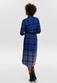 Niebieska sukienka w kratkę z długim rękawem i czarnymi akcentami, dopasowana w talii, z frędzlami u dołu, zestawiona z czarnymi botkami za kostkę. Zaprezentowany widok z tyłu.