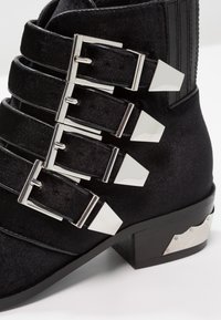 Stivaletto nero con più fibbie metalliche, tessuto testurizzato e tacco quadrato con un accento argentato.