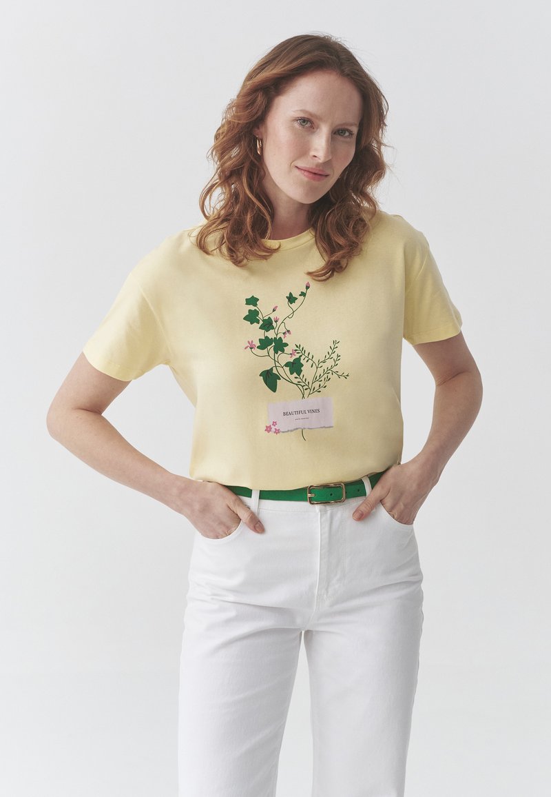 Camiseta amarilla de manga corta con un diseño floral que incluye hojas verdes y flores rosas. Combinada con pantalones blancos y un cinturón verde.