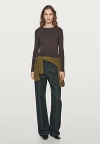 Mørkebrun langærmet skjorte, løse mørke denimbukser, olivengrøn sweater bundet om taljen, mørke sko, neutral baggrund.