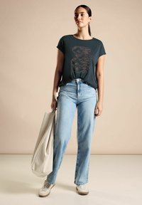 T-shirt verde scuro a maniche corte con design testuale, abbinato a jeans a vita alta blu chiaro e sneakers bianche, mentre tiene una borsa tote beige.