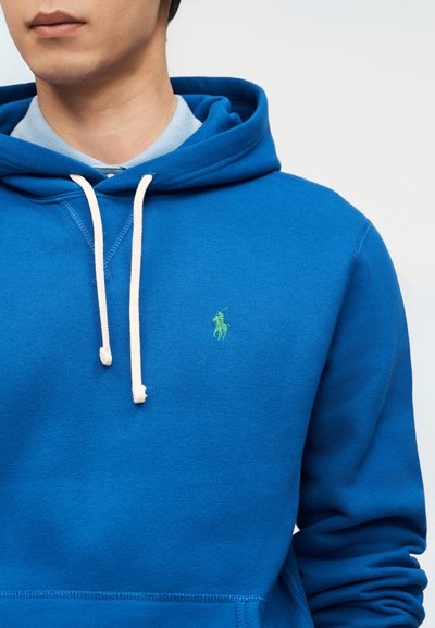 Νεαρός άνδρας φορώντας φωτεινό μπλε φούτερ με λευκά κορδόνια και ανοιχτό μπλε πουκάμισο με γιακά από μέσα, με ορατό πράσινο λογότυπο παίκτη πόλο στο στήθος.