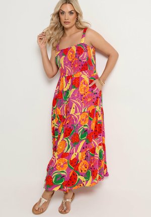 Kvinde iført farverig maxikjole med frugtmønster, poserer med den ene hånd på hoften og sandaler på fødderne mod en ensfarvet baggrund.