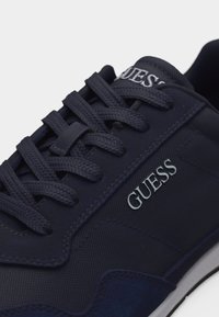 Baskets Guess bleu marine en tissu texturé, lacets épais, détails cousus, logo blanc sur le côté et sur la languette.
