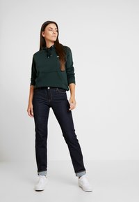 Sudadera verde oscura con bolsillo frontal, vaqueros oscuros ajustados y zapatillas blancas. Textura suave, diseño casual y puños doblados en los vaqueros.