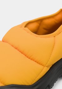 Botte slip-on rembourrée jaune avec une surface texturée et une semelle en caoutchouc noir. Présente un bout arrondi et des détails cousus.
