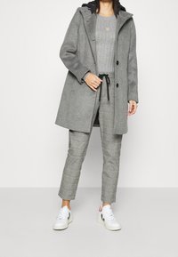 Cappotto in lana grigio chiaro con cappuccio, due bottoni e tasche laterali. Abbinato a un maglione grigio a coste e pantaloni scozzesi. Sneakers bianche.