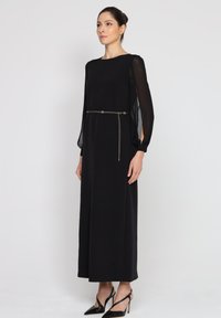 Czarna maxi sukienka z długimi, przezroczystymi rękawami, podkreślona cienkim paskiem oraz łańcuszkowym detalem w talii. Gładki materiał i luźny krój.