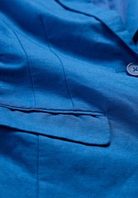 Niebieska lniana marynarka z kieszenią na lewym piersi, subtelną teksturą i tonowymi detalami szycia. Widoczne zapięcie na guziki w pobliżu kołnierza.