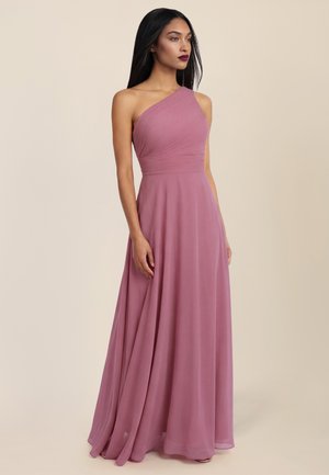 Frau mit langen dunklen Haaren, die ein bodenlanges, einseitig schulterfreies Abendkleid in Mauve trägt und vor einem schlichten beige Hintergrund steht.