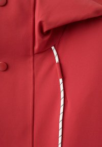 Tissu rouge avec deux boutons visibles à gauche et un cordon blanc avec des tirets noirs passant à travers un petit porte-tissu à droite.