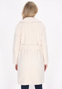 Cappotto soffice di colore crema con vita con cintura, maniche lunghe e colletto; presenta un tessuto testurizzato e morbido. Vista da dietro mostrata.