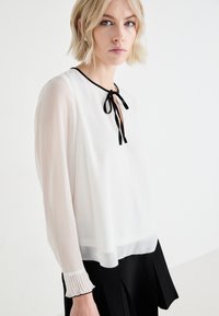 Blouse blanche avec cravate noire, manches longues, encolure ronde, détails de poignets plissés, tissu fluide, ourlet noir contrastant, design moderne et léger.