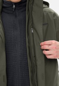 Olijfgroene buitenjas met een gladde textuur, gelaagd over een zwarte fleece met structuur, met ritsen en klittenbandsluitingen.