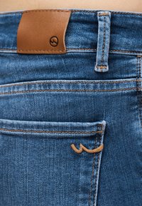 Κοντινή λήψη πίσω τσέπης μπλε τζιν με δερμάτινο ταμπελάκι σε ταμπά απόχρωση και λεπτομέρεια κεντήματος σε ταμπά κοντά στην άκρη της τσέπης.