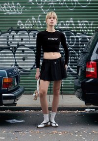Black long-sleeve crop top with logo, black pleated skirt, white ankle socks, and black shoes with white accents. Plush toy held in left hand.
