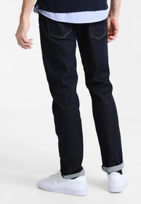 Dark blue denim jeans with a slim fit, featuring rolled cuffs and standard back pockets. Worn with white sneakers and a layered shirt.
